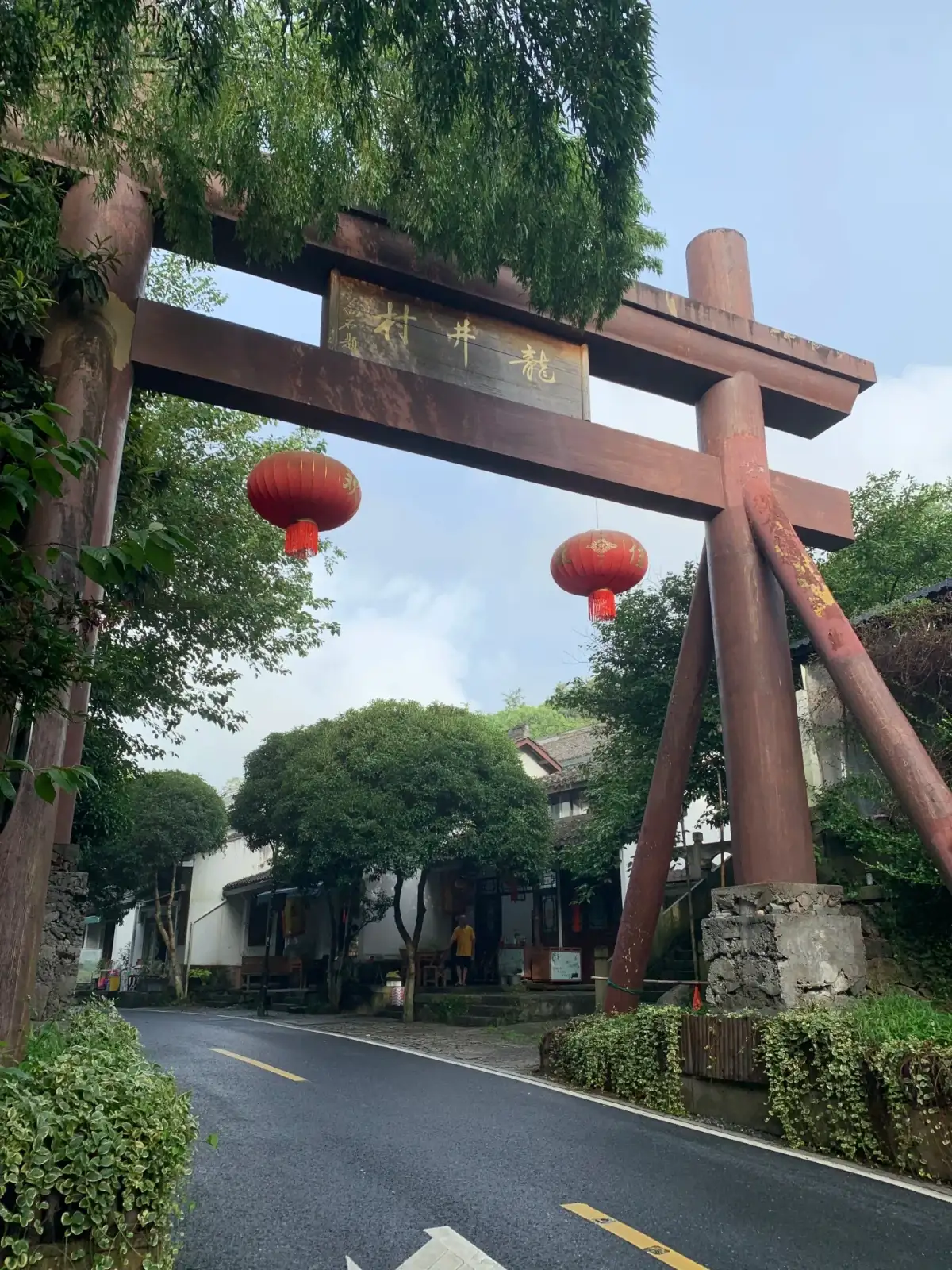 A wooden gate spans the road, with red lanterns hanging by the entrance and dense trees lining both sides.