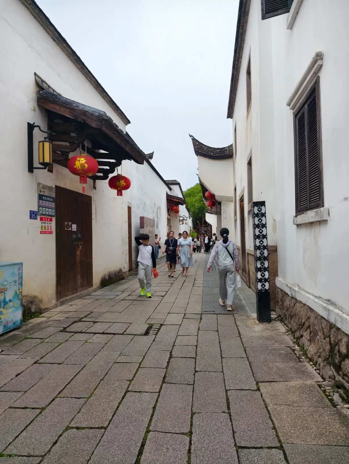 Red lanterns hang in front of the door of a narrow alley with white walls, and in the distance, someone is walking along the stone-paved path.
