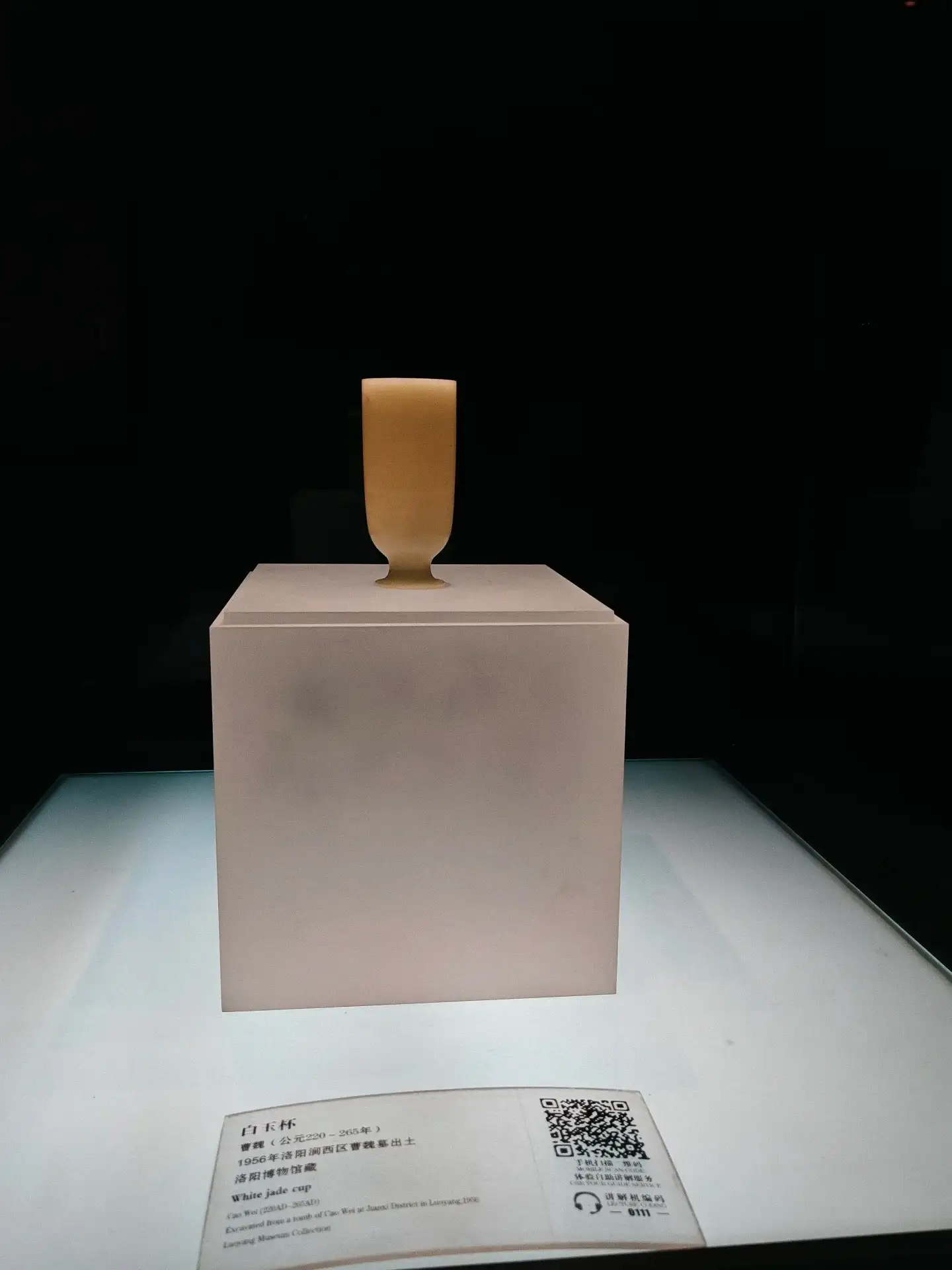 In the dimly lit exhibition hall, a light-colored, cup-shaped artifact stands on a high display platform.