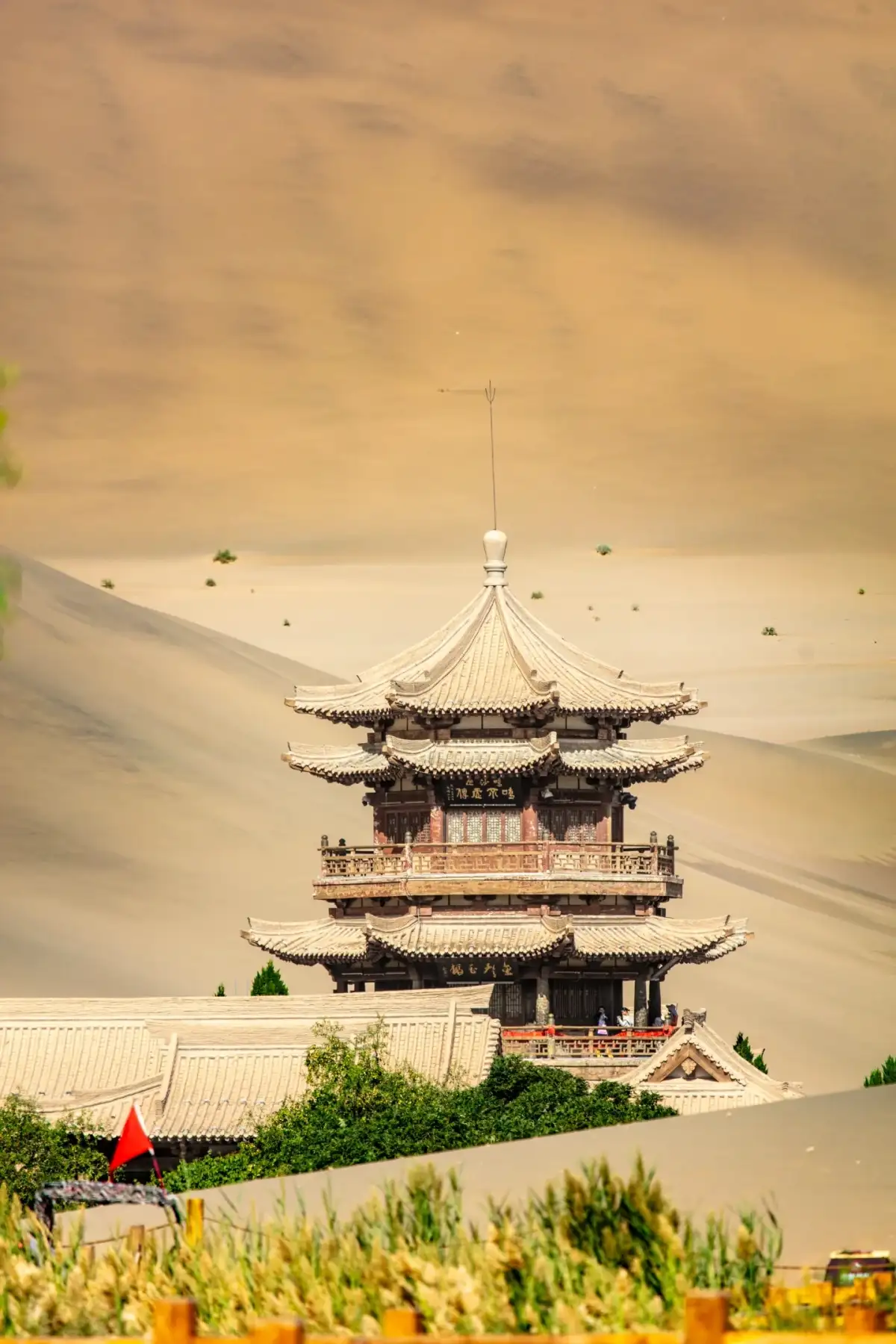 多层木构楼阁立在沙丘前,前景是绿树和较低的瓦屋顶。