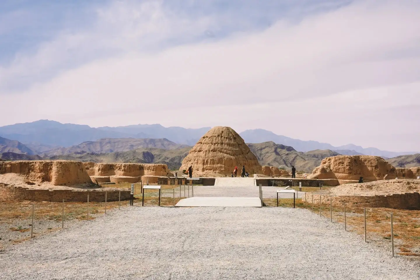 A gravel path leads to the earthen ruins, where a central, elevated mound stands in contrast to the distant mountain range.