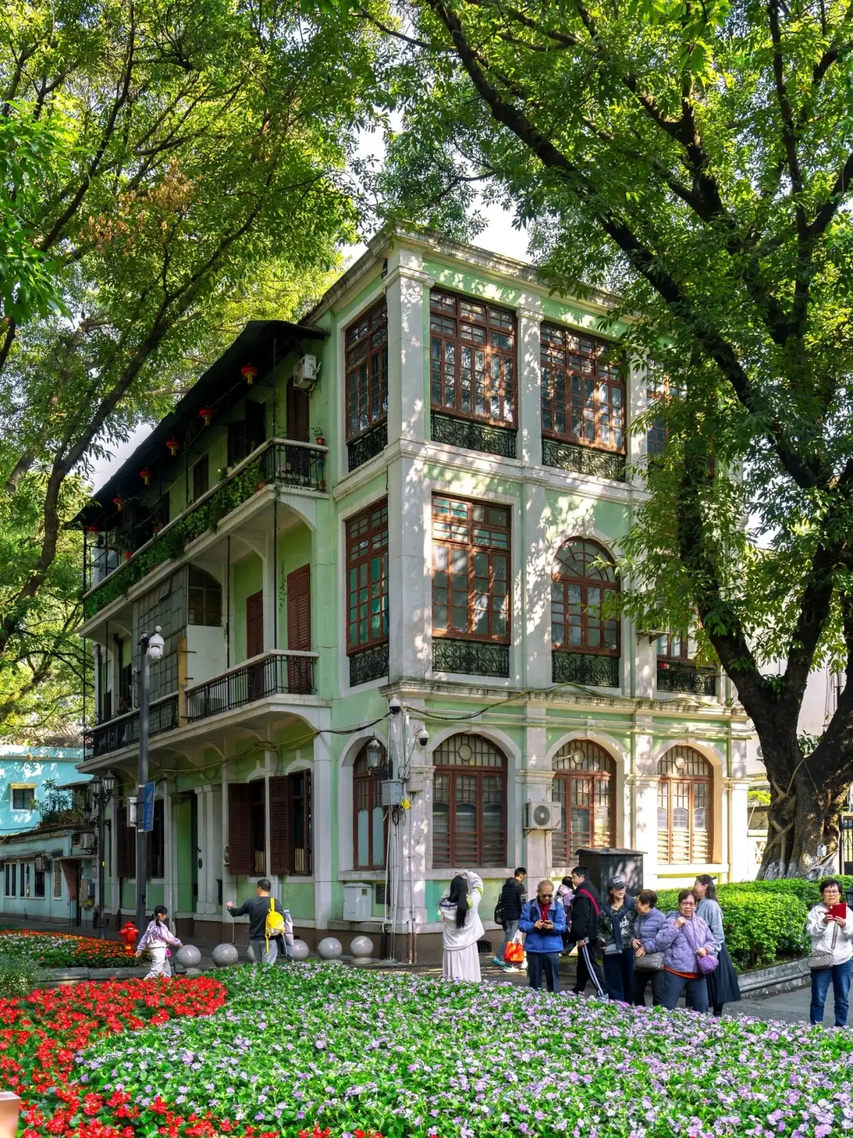 绿色装饰立面转角旁长着大树,前景花坛与远处游客构成街景。