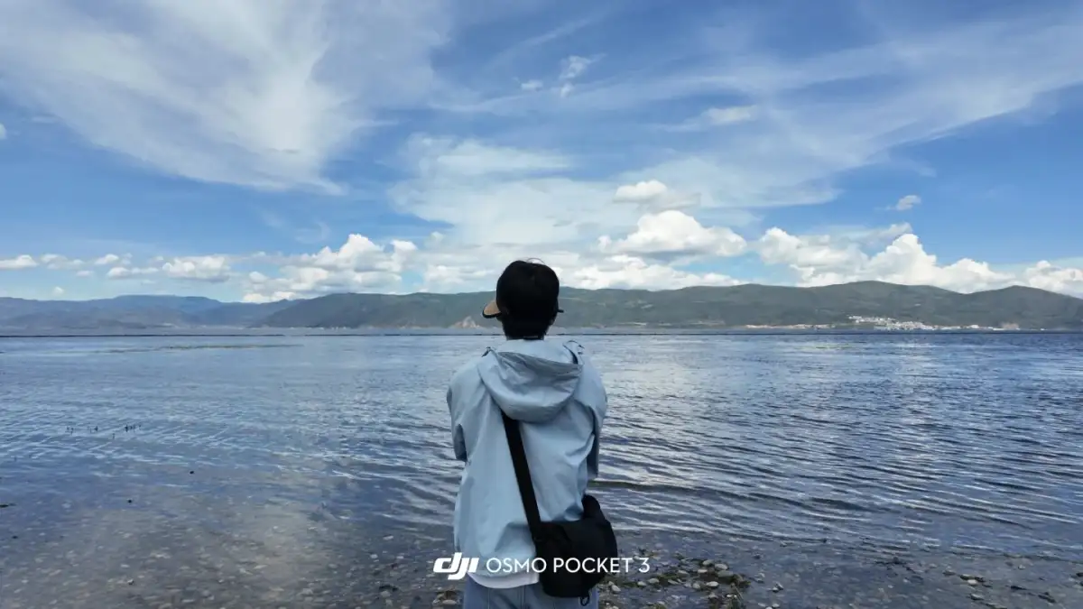 A traveler with their back to the camera faces the open lake, where the distant mountains and white clouds merge into a single line.