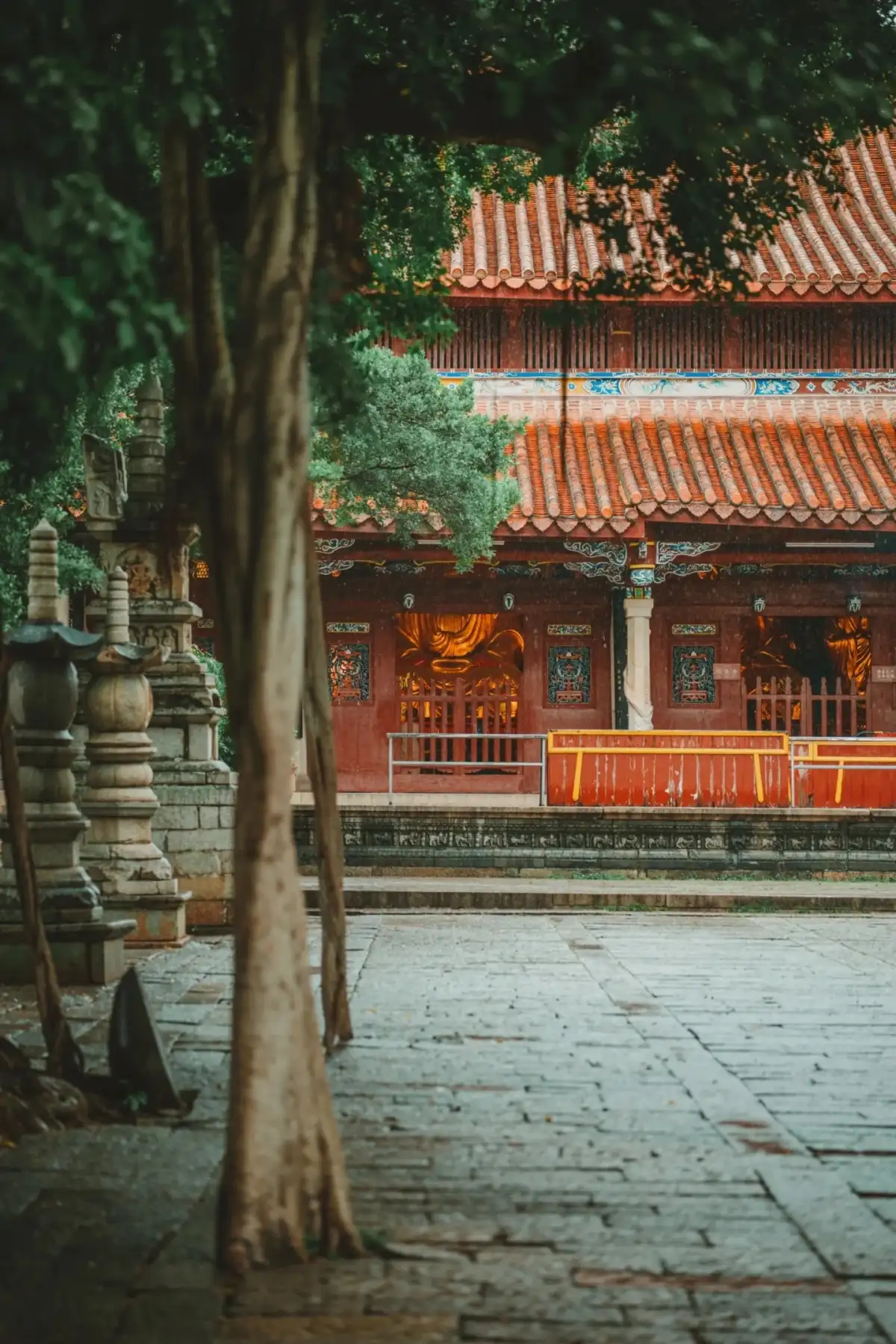 The stone-paved courtyard in front of the large tree glistens with a dewy sheen, flanked by red halls and stone lanterns.