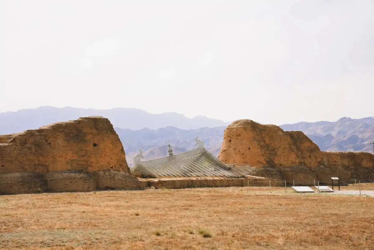 a gray roof peeking out between two sections of earthen wall, set against a backdrop of distant mountains and an open sky.
