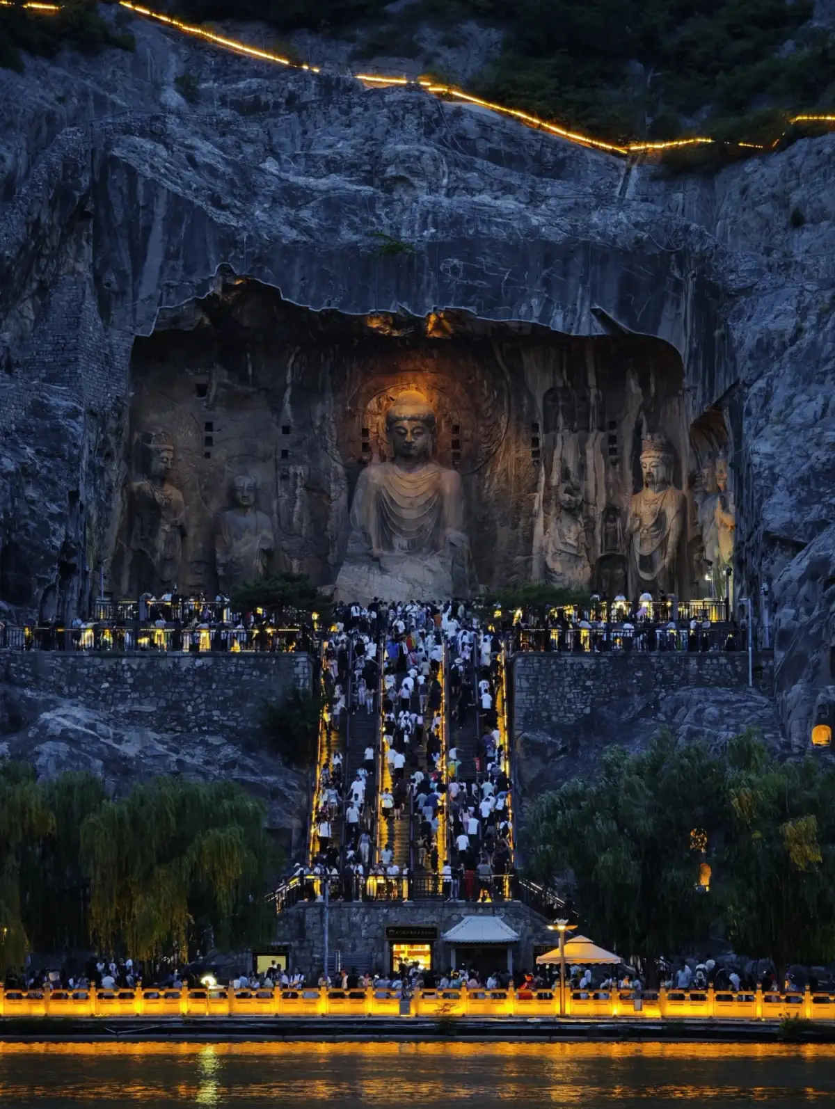 The cliff-side grottoes are illuminated by lights in the night, and the visitors on the steps blend with the play of light and shadow on the river’s surface.
