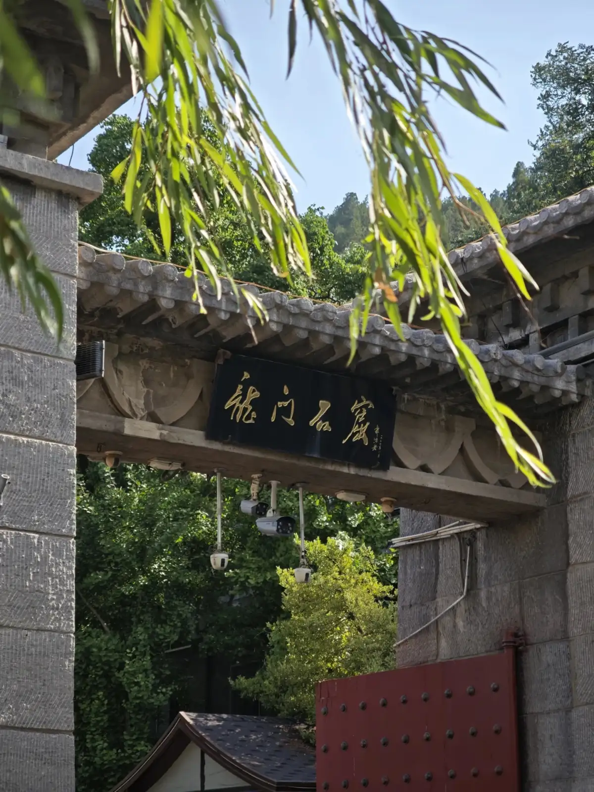 Weeping willows shade the gray-tiled gatehouse; beneath the gate, red door panels and a black-backed plaque with calligraphy are visible.