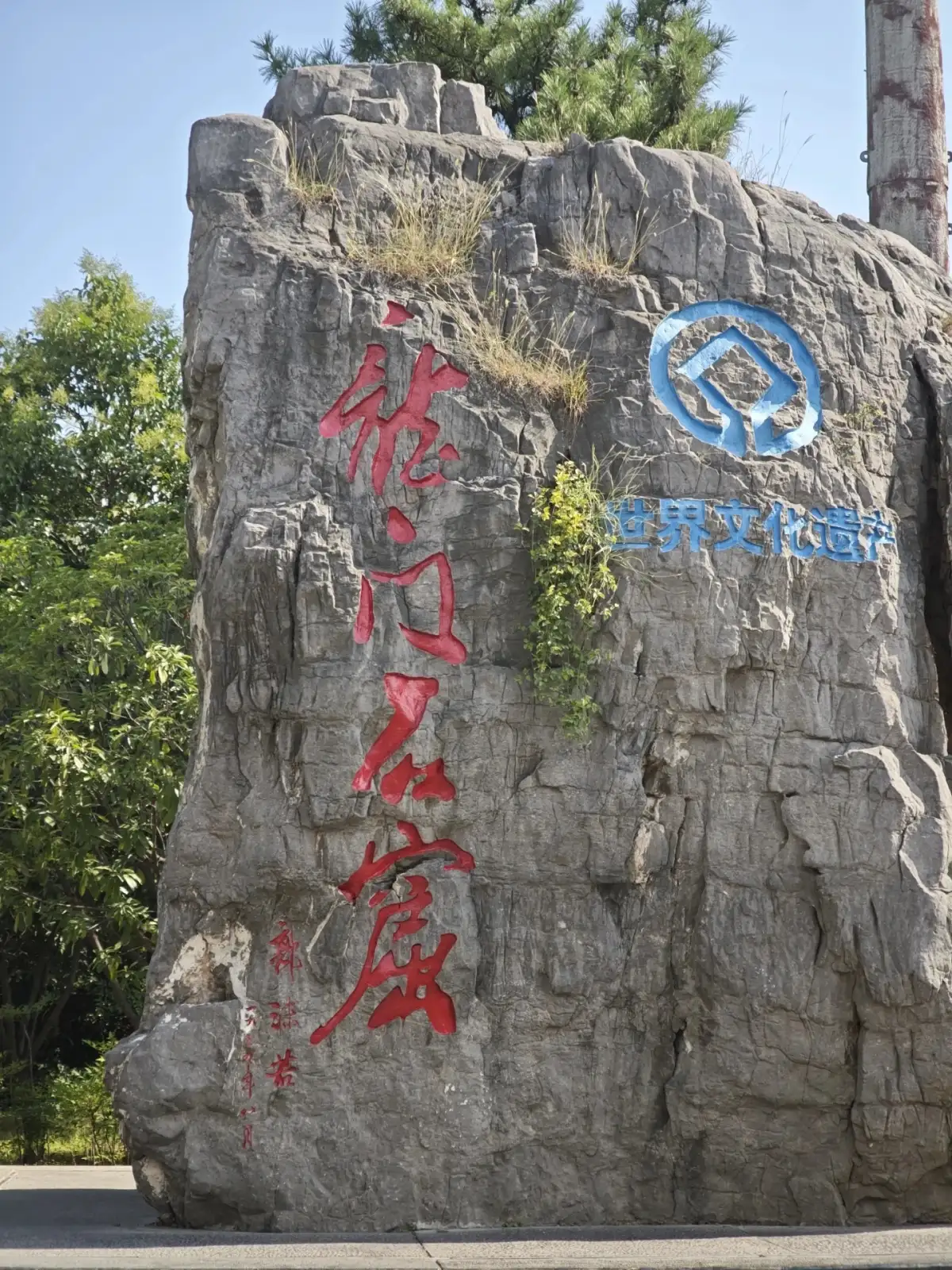 The words “Longmen Grottoes” are carved in red on a large stone block, with a blue sign next to it.